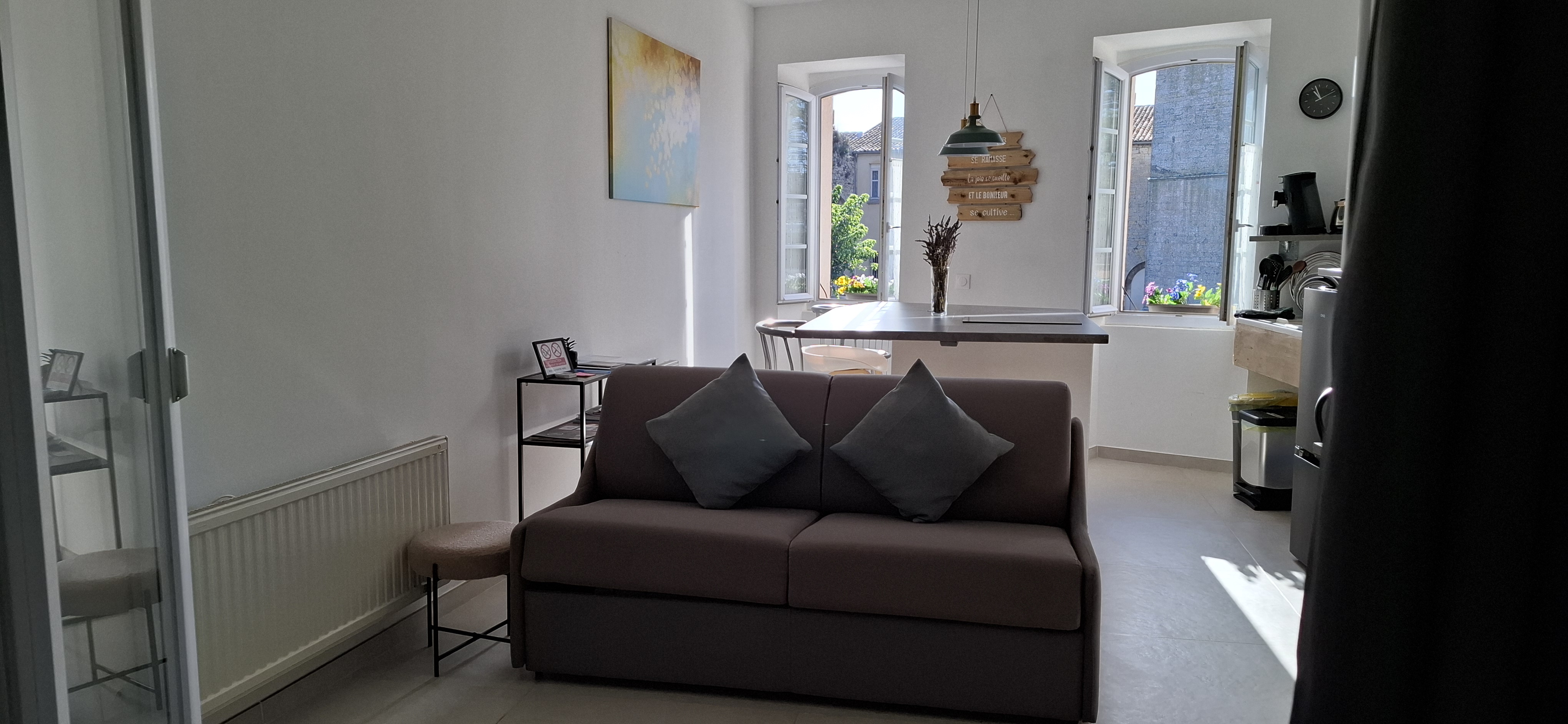 A room with a couch and a table, featuring an artistic painting on a white wall and a window in the background.